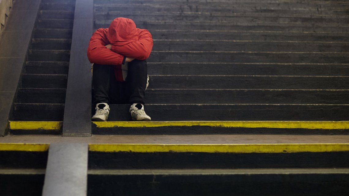 Person in roter Winterjacke sitzt zusammengekauert auf einer öffentlichen Treppe. Das Gesicht ist in den auf den Knies verschränkten Armen verborgen.