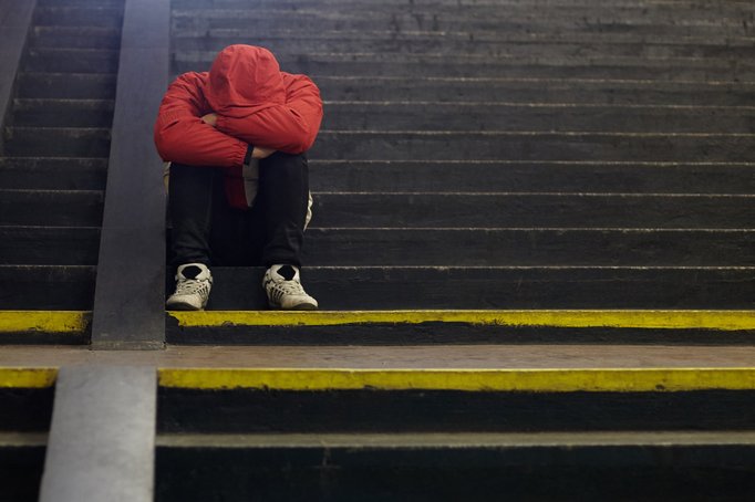 Person in roter Winterjacke sitzt zusammengekauert auf einer öffentlichen Treppe. Das Gesicht ist in den auf den Knies verschränkten Armen verborgen.